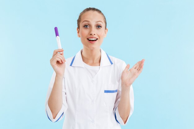 Beautiful excited blonde woman doctor wearing uniform standing isolated over blue wall, showing pregnancy test