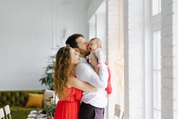 A beautiful family with a baby in their arms stand near the window in the living room