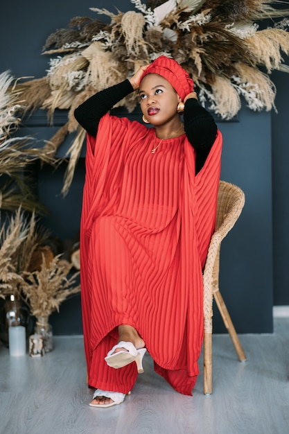 Beautiful fashion model wearing red traditional african outfit and head shawl, earrings and bright makeup, sitting on the wicker chair on dark studio background with boho decor