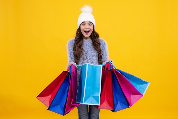 Beautiful fashion teenager child girl in winter warm hat with shopping bags on yellow background