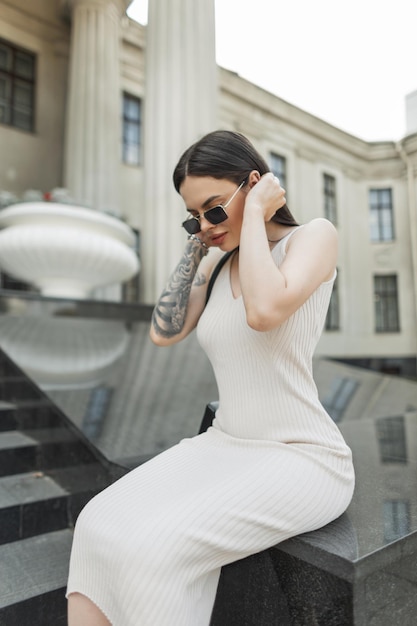 Photo beautiful fashion trendy urban girl with vintage sunglasses in a fashion beige dress with a tattoo on her arm sits near a column building on the street and straightens her hair pretty hipster woman