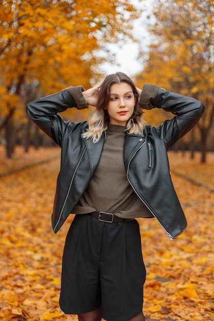 Beautiful fashion young girl in stylish clothes with a leather jacket sweater and shorts walks in the autumn golden park Beauty style and fashion