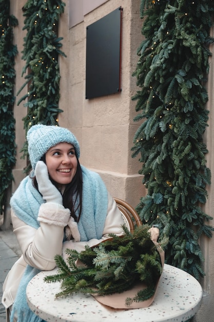 Beautiful fashionable woman sitting on the chair at outdoors cafe with christmas tree bouquet