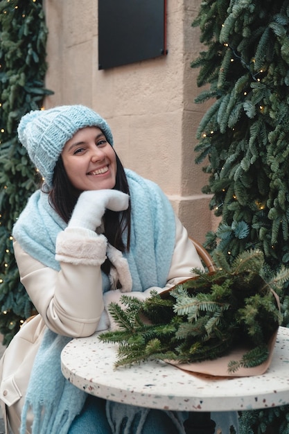 Beautiful fashionable woman sitting on the chair at outdoors cafe with christmas tree bouquet