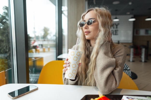 Beautiful fashionable woman with a blond curly hairstyle with vintage sunglasses in a beige sweater sits in a cafe and drinks a cola near the window