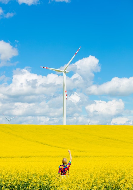 Beautiful female in 90s stylish shirt in rapeseed field with wind turbine on background