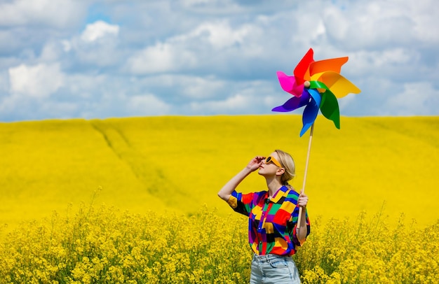 Beautiful female in 90s stylish shirt with pinwheel in rapeseed field