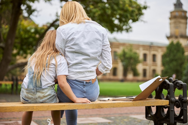 beautiful female in casual clothes with her daughter in denim romper spending time together in the park