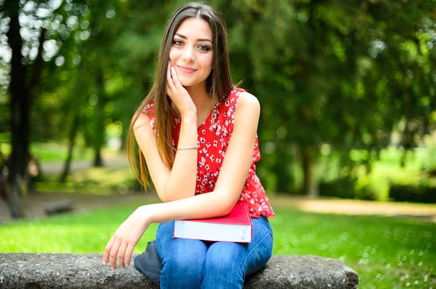 Beautiful female college sitting on a bench in the park and holding a book on her knees