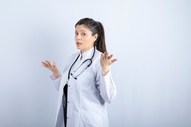 Beautiful female doctor in white coat posing with stethoscope over white wall.