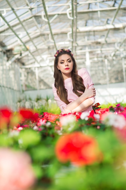 Beautiful female model dressed in a long pink dress poses among the many flowers in the greenhouse