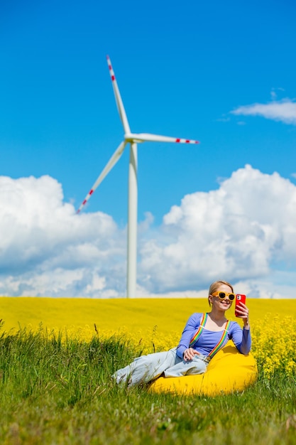 Beautiful female in rainbow LGBT suspenders for pants and mobile phone is sitting in bean chair in rapeseed field