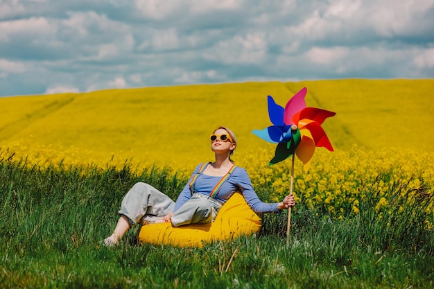 Beautiful female in rainbow LGBT suspenders for pants sit in bean bag with pinwheel in rapeseed field