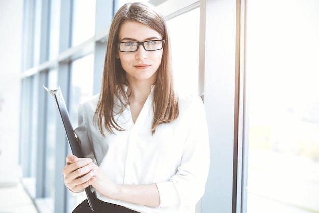 Photo beautiful female specialist with clipboard standing in sunny office and smiling charmingly business