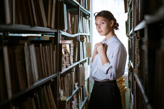 Beautiful female student in a white shirt stands between the rows in the library, bookshelves worth of books. Dark photo