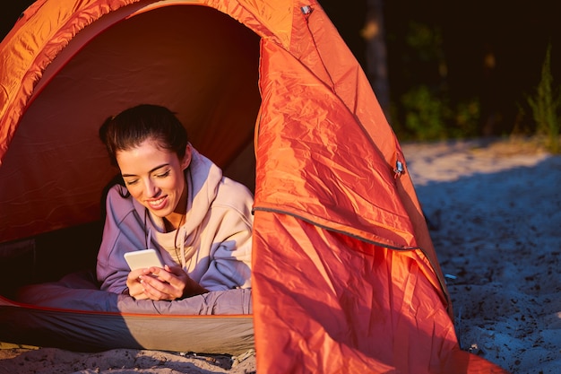 Beautiful female traveler texting message on smartphone and smiling while resting in touristic tent