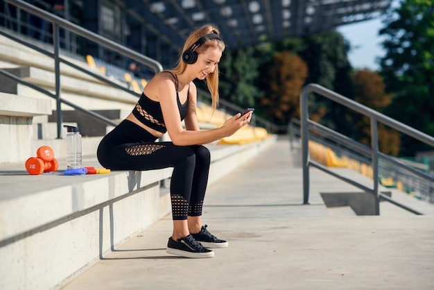 A beautiful fitness girl in grey sportswear uses smartphone and listens to music at the stadium after workout. Sports and healthy concept.