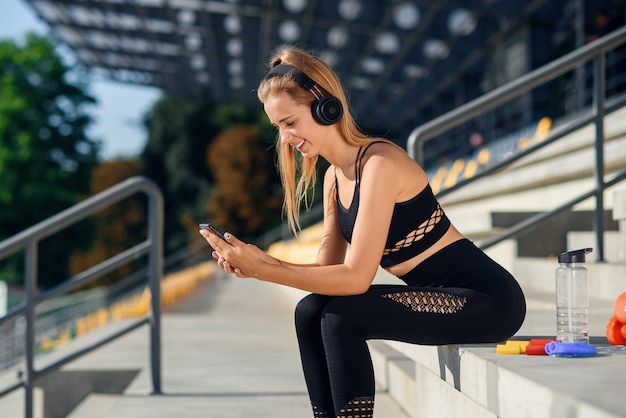 A beautiful fitness girl in grey sportswear uses smartphone and listens to music at the stadium after workout.