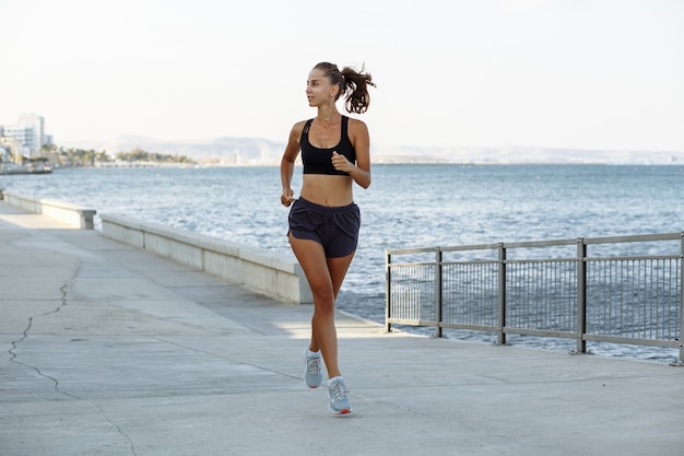 Beautiful fitness girl in shorts and sports top running on the waterfront by the sea in sunny weather