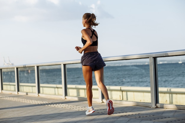 Beautiful fitness girl in shorts and sports top running on the waterfront by the sea in sunny weather