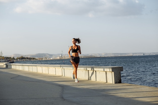 Beautiful fitness girl in shorts and sports top runs on the waterfront by the sea during sunset