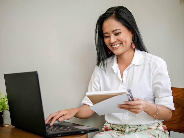 Beautiful freelancer holding a book in hand, typing on laptop 