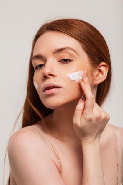 Beautiful fresh young redhead woman with red hair applies a cream on her face and does a skin moisturizing Beauty freshness and skin care