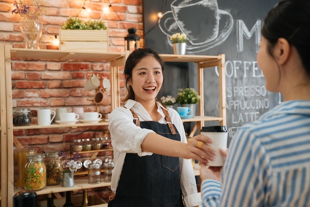 Beautiful friendly female barista giving coffee disposable cup to customer at counter of coffee shop. small startup business owner concept. office lady is regular client buy breakfast every morning