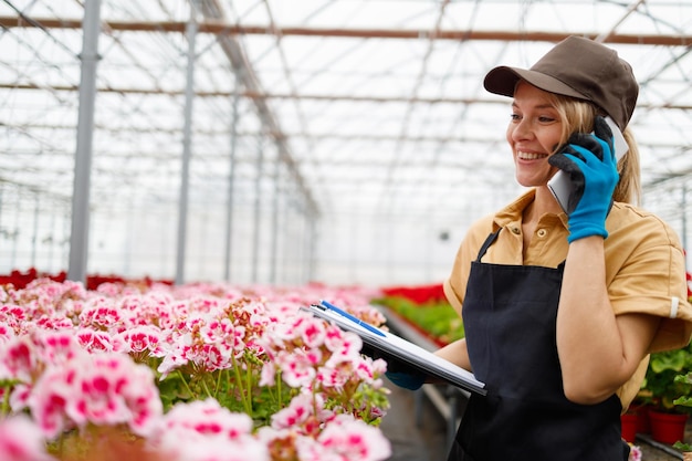 Beautiful gardener woman talking on the phone with a client and makes notes in greenhouse