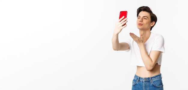 Beautiful gay man in crop top sending air kiss at phone camera taking selfie or video chat on smartphone standing over white background