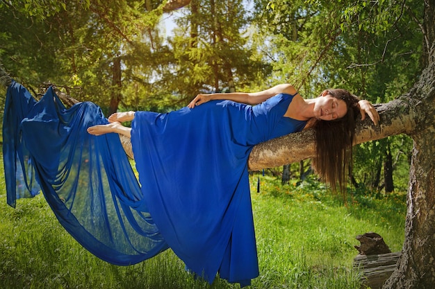 A beautiful girl of Asian appearance with long hair in a blue dress lies on a tree branch in Altai. Tourism concept.