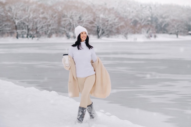 A beautiful girl in a beige cardigan and a white hat with a glass of tea enjoys a snowy embankment by the lake