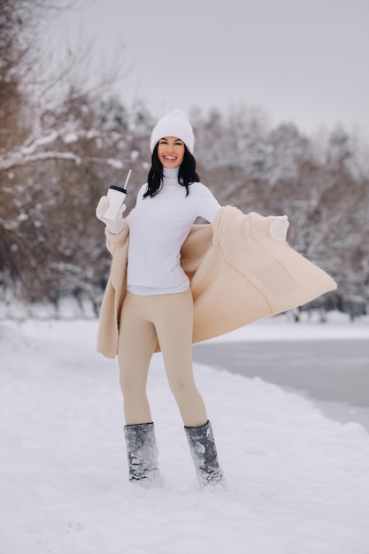 A beautiful girl in a beige cardigan and a white hat with a glass of tea enjoys a snowy embankment by the lake