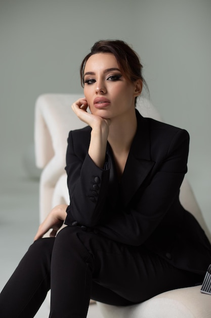 A beautiful girl in a black business suit is sitting in a chair on a gray background