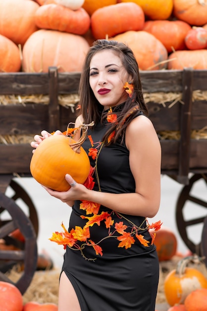 Beautiful girl in a black dress decorated with yellow leaves poses near a wheelbarrow filled with various pumpkins He holds a pumpkin in his hands Halloween pumpkin Pumpkin decor