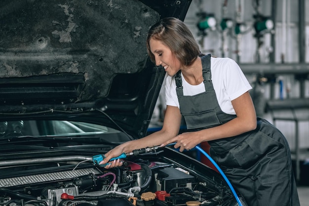 A beautiful girl in a black jumpsuit and a white t-shirt is smiling, checking the oil level in a black car in the garage.