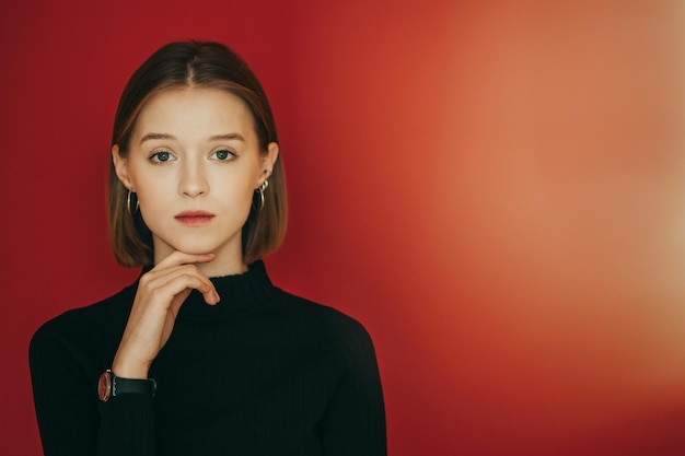 Beautiful girl in a black sweater stands in the studio on a red background looks into the camera