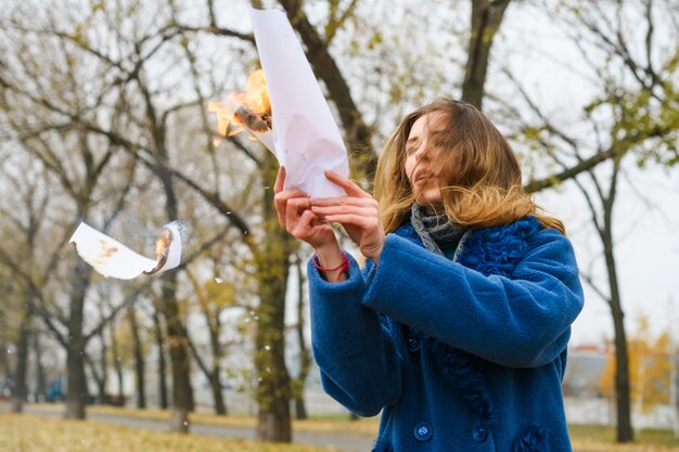 Beautiful girl in blue coat make ritual with burning white paper sheet at nature background, fighting depression