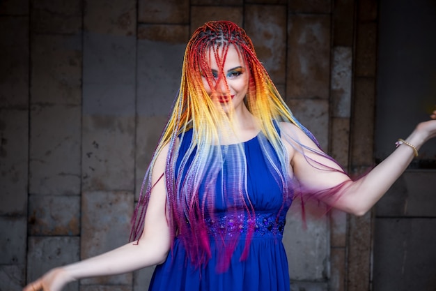 A beautiful girl in a bright blue dress with rainbow African braids and unusual glitter makeup. Spins and laughs against the background of an old large building on the warm streets of the spring city.