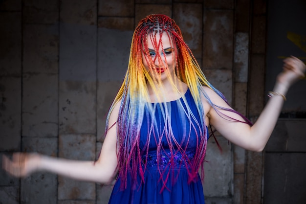 A beautiful girl in a bright blue dress with rainbow African braids and unusual glitter makeup. Spins and laughs against the background of an old large building on the warm streets of the spring city.