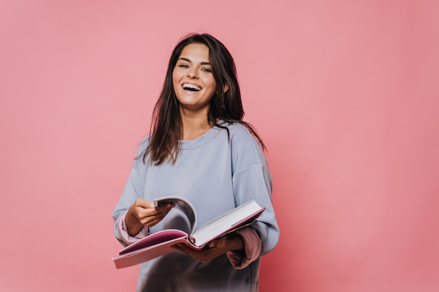 Beautiful girl in casual sweater with loose hair leafs open book on pink background and laughing. Enjoying the new book. Happy people concept.