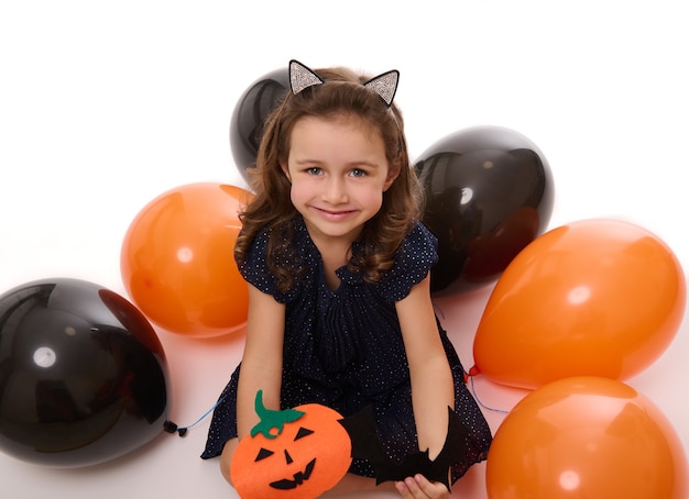 Beautiful girl child smiles with toothy smile , playing with homemade felt-cut pumpkin on the white background with lying down inflated black and orange colored balloons. Halloween concept, copy space