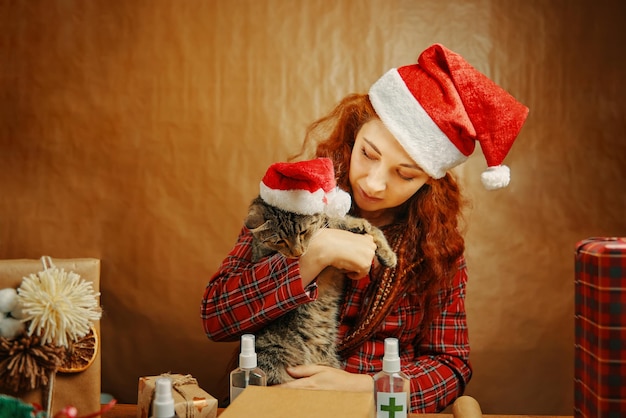 Beautiful girl in christmas hat with cat in santa claus hat near table with gift boxes and sanitizer...