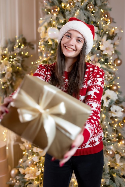 Beautiful girl in a Christmas sweater and a red Santa Claus hat sits near a luxurious Christmas tree Smiling Holds a gift in his hands Celebration of new year christmas