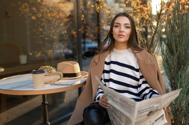 Beautiful girl in a coat reads a newspaper and smiles while relaxing in an outdoor cafe