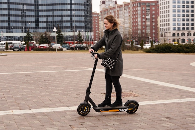 Beautiful girl in a coat rides electric scooter around the city