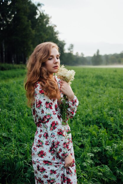 A beautiful girl in a colorful dress stands in the evening on a meadow against a background of fog. 