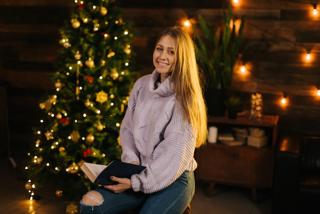 Beautiful girl in cozy sweater posing with a book in her hand against the background of a Christmas tree and wall with a festive garland. Nice girl looking at the camera.