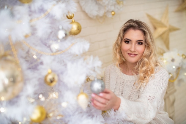 Beautiful girl decorating Christmas tree. a young smiling woman prepares a Christmas tree for the holiday. blonde in a light blouse. lush white Christmas tree with Golden balls