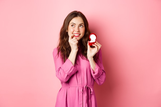 Beautiful girl dream of her wedding day, looking thoughtful up, showing engagement ring in red box and smiling, standing over pink wall.